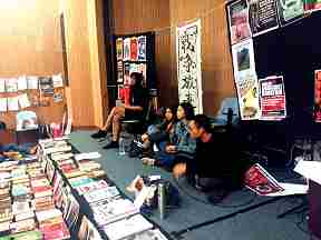 Collective members from a vegan restaurant in a working class community with a “pay what you feel is ight” free/autonomoous pricing operation presenting at the Hong Kong book fair. Image shows 4 people sitting on a low stage, many publications and posters in Chinese and English are visible.