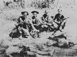 Photo shows four Marines sitting in a field. In front of them are various body parts, including heads. They are all heavily armed.