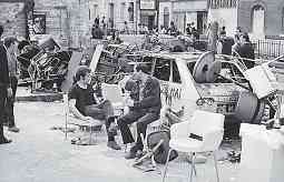 Photo shows two seated men in discussion amid piles of furniture and other objects.
