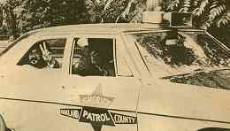 Photo of John Sinclair giving peace sign in back seat of police car.