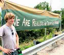 Photo of a smiling young man standing near a large banner that reads “We are the realists: We demand the impossible."
