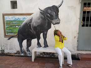 Photo shows a brown-skinned woman sitting on a bench. On a wall behind her is a life-size painting of a bull, appearing as if its forehooves are on the bench. Mbeke pretends to be afraid.
