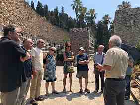 Photo showing a small group of people standing in a circle in an outdoor setting, surrounded by stone walls, in full sun.