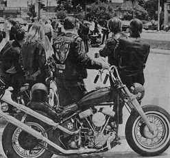 Photo shows a group of young people standing in a lot with several motorcycles.
