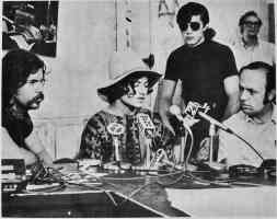 photo shows three people sitting at a table; microphones from several radio and/or TV stations are visible.