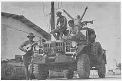 Photo shows 4 GIs on and around a military vehicle. One is playing a guitar while others seem to be in relaxation mode.