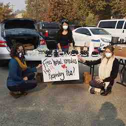 Three women wearing protective face masks share herbal remedies from a table in a parking lot.