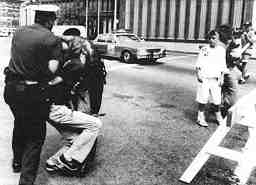 Photo shows an officer exerting force on the arm and neck of a protester as children look on.