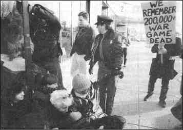 photo shows women sitting on the ground. A sign reads, "We remember 200,000 war game dead."