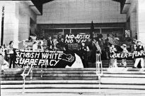 Photo shows a large group of people in front of a building. Signs read slogans such as "Smash white supremacy."