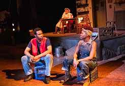 Photo shows two seated young actors engaged in conversation as an older woman looks on from a distance.