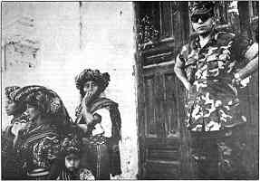 photo shows 3 indigenous women and a small girl with concerned or fearful expressions. Behind them a scowling U.S. soldier in camouflage looks on.