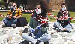 Photo shows three masked people sitting cross-legged on the ground, one with musical instruments two holding microphones. Caution tape surrounds and connects them.