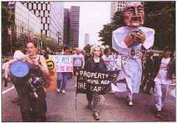 Photo shows protesters in the street with musical instruments and a giant puppet. A sign reads, "Property is a crime against the Earth."