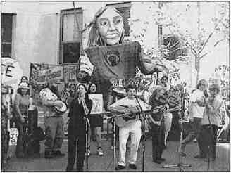 Photo shows protesters with signs, musicians performing and a large puppet in an outdoor setting