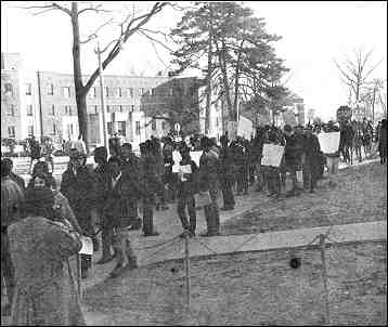 Photo shows a crowd assembled on a sidewalk. Many people are holding signs, which are not legible.