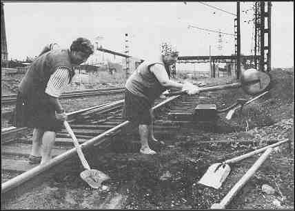 Photo shows two women shoveling gravel and debris along a railroad track.