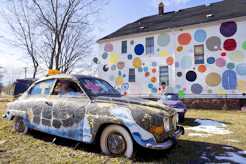 Photo shows an artistically painted car next to a house painted with large dots of many hues.