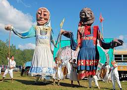 A color photo shows 2 giant female figure puppets being manipulated by the use of long poles. The figures appear to be about 15 feet high.