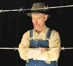 Photo depicts Norman Nawrocki in coveralls and hat standing behind two strands of barbed wire. With arms folded, he has a serious expression on his face.