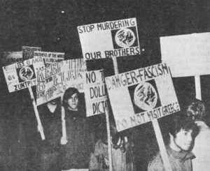 Photo shows a small crowd of demonstrators. Signs include "Danger: Fascism, Do Not Visit Greece" and "Stop Murdering Our Brothers"