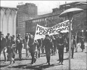 A group of young people is shown marching in the street. They are carrying a large banner that reads, "Revolutionary Contingent."