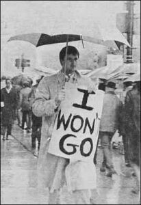 Photo shows a crowd of people protesting in the rain. A young man holds a hand-lettered sign reading, "I won't go."