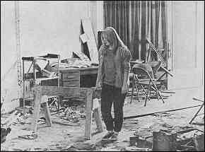 Photo shows a young woman somberly surveying a severely damaged office. Broken furniture and piles of torn paper goods are visible.