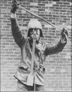 Photo shows a man wearing a gas mask, crash helmet and protective jacket. A brick wall is in the background.