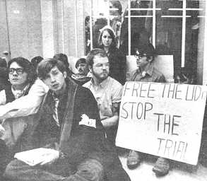 Photo shows a group of ten or more young people sitting on a floor. A partially-visible sign reads "Free the UD ... Stop the trial."