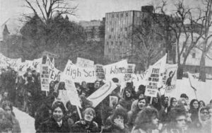 Photo shows a crowd, mostly women, many with signs, in an urban setting.
